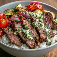 Sliced, juicy grilled steak bowl over fluffy rice and colorful roasted vegetables, drizzled with bright green homemade chimichurri sauce for dinner.